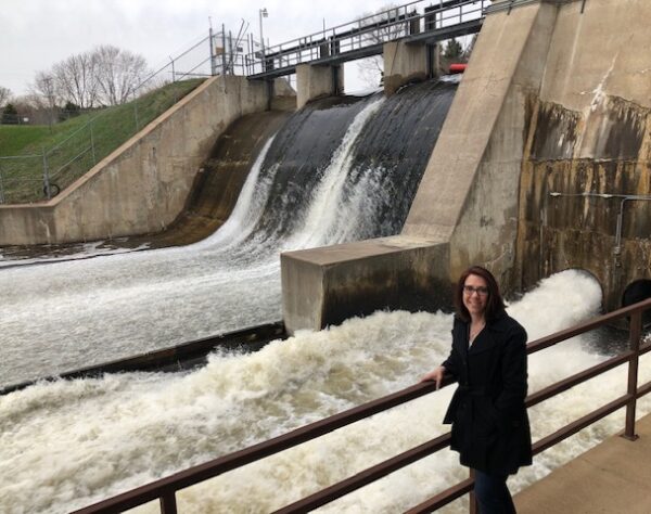 A brown-haired woman stands in front of a river dam with rushing water flowing behind the railing she is leaning on.