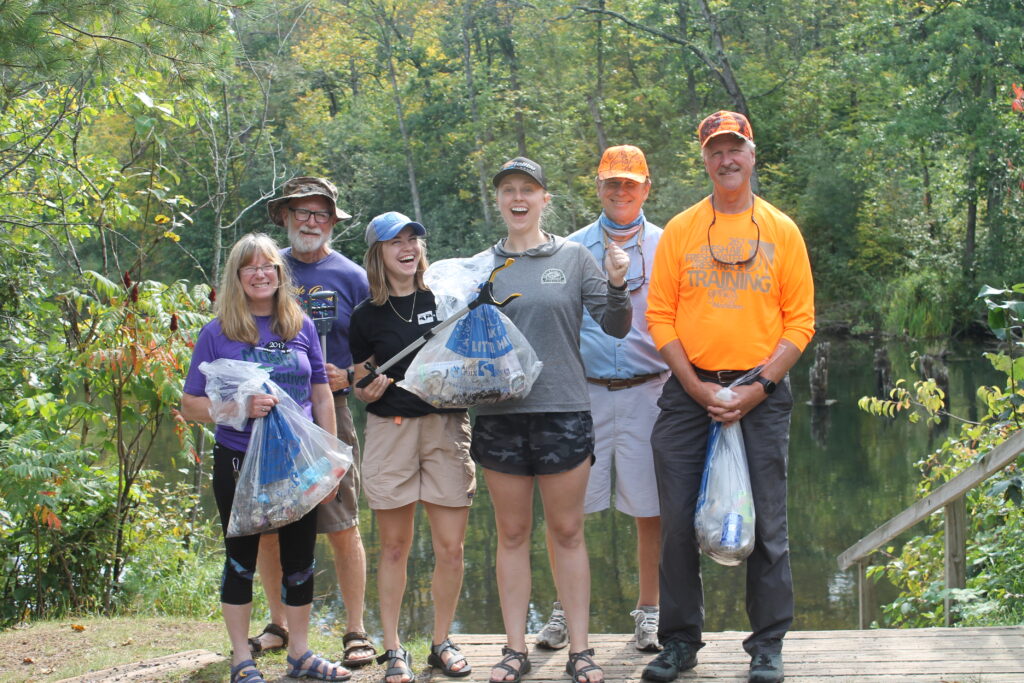 A group of volunteers stand near a river. They are holding trash grabbers and trash bags.