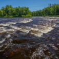 A blue sky frames a photo of the St. Croix River. Brown water rushes over rocks, creating a small series of rapids