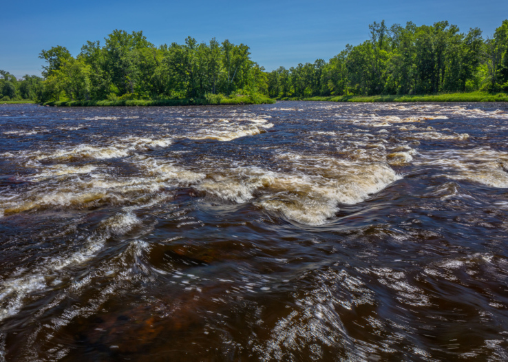 A blue sky frames a photo of the St. Croix River. Brown water rushes over rocks, creating a small series of rapids