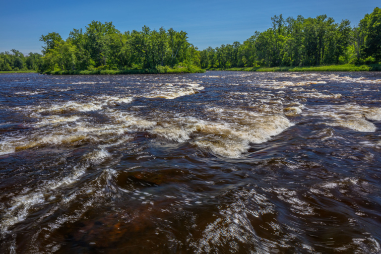 A blue sky frames a photo of the St. Croix River. Brown water rushes over rocks, creating a small series of rapids