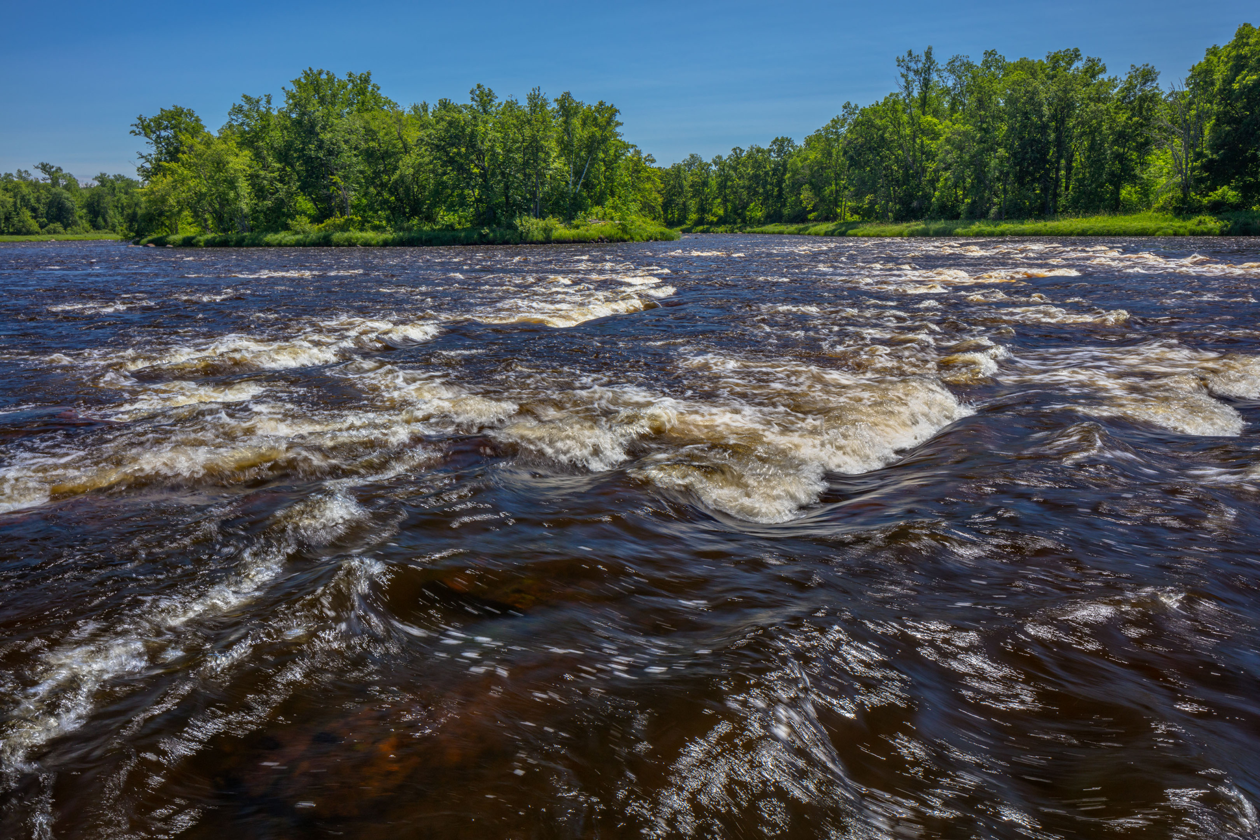 A blue sky frames a photo of the St. Croix River. Brown water rushes over rocks, creating a small series of rapids