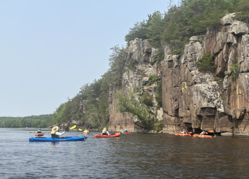 A group of kayakers on the St. Croix River next to a tall cliff face.