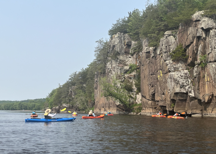 A group of kayakers on the St. Croix River next to a tall cliff face.