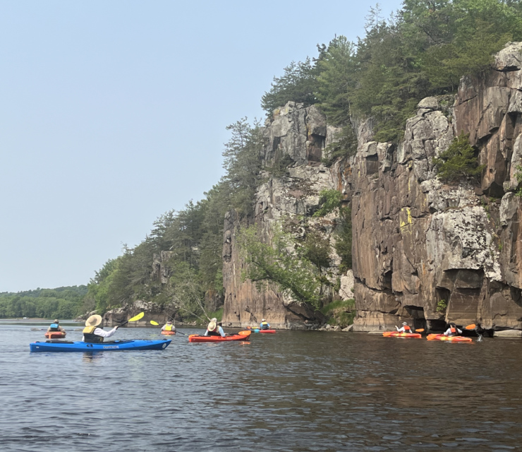 A group of kayakers on the St. Croix River next to a tall cliff face.