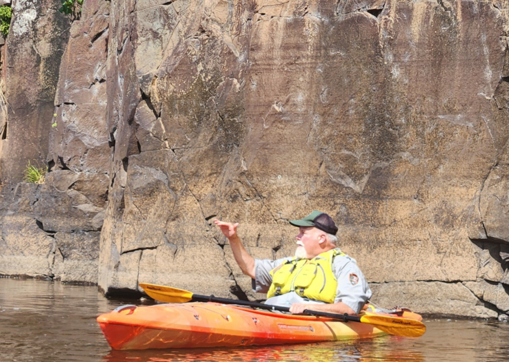 A park ranger in an orange kayak in front of a large rock face on the river