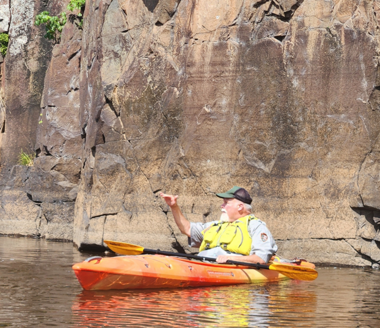 A park ranger in an orange kayak in front of a large rock face on the river