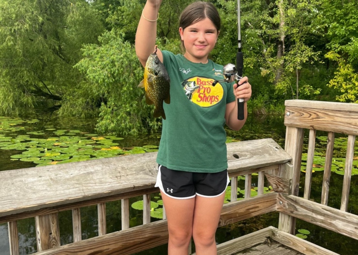 A young girl stands holding up her fishing rod and fish